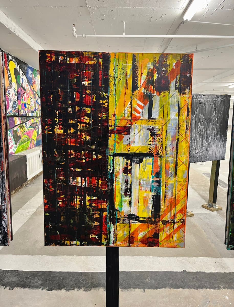 A photograph of a large, abstract painting displayed on a stand in a gallery or studio space. The artwork features a split composition with dark, textured vertical strokes on the left and bright yellow, orange, and white geometric patterns on the right.