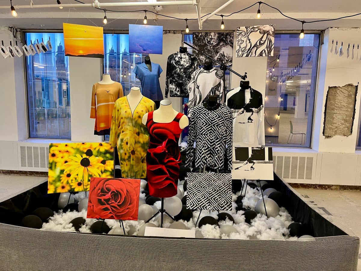 An art installation featuring mannequins dressed in patterned clothing and several framed paintings displayed on easels within a large, fabric-lined pit filled with white and black balloons. The scene is set in a room with large windows and string lights.