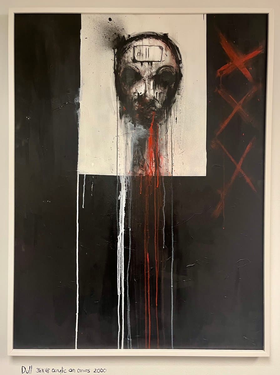 A framed abstract painting featuring a dark, mask-like face with a white rectangle on its forehead and red drips running down its center, set against a black and white background with red X marks on the right. Below the frame, handwritten text on the wall reads 'Dull 36x48 acrylic on canvas 2000'.