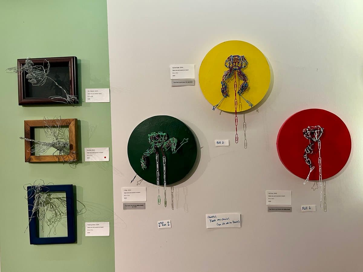 A gallery wall displaying six mixed-media art pieces: three framed wire sculptures on the left and three circular canvases with wire and paperclip figures on the right. Small informational placards are mounted next to each artwork, with one handwritten sign reading "(squeaky) Yank my chain! (yes it's ok to touch)".