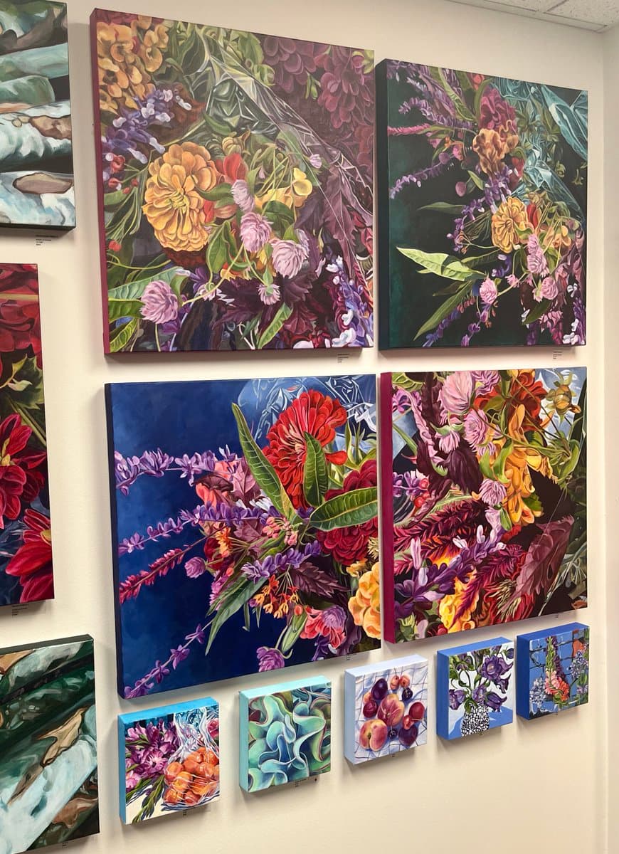 A gallery wall displaying a collection of vibrant, colorful paintings of flowers and botanical subjects in various sizes. The artworks feature rich, saturated hues and detailed brushwork, arranged in a grid-like pattern on a white wall.