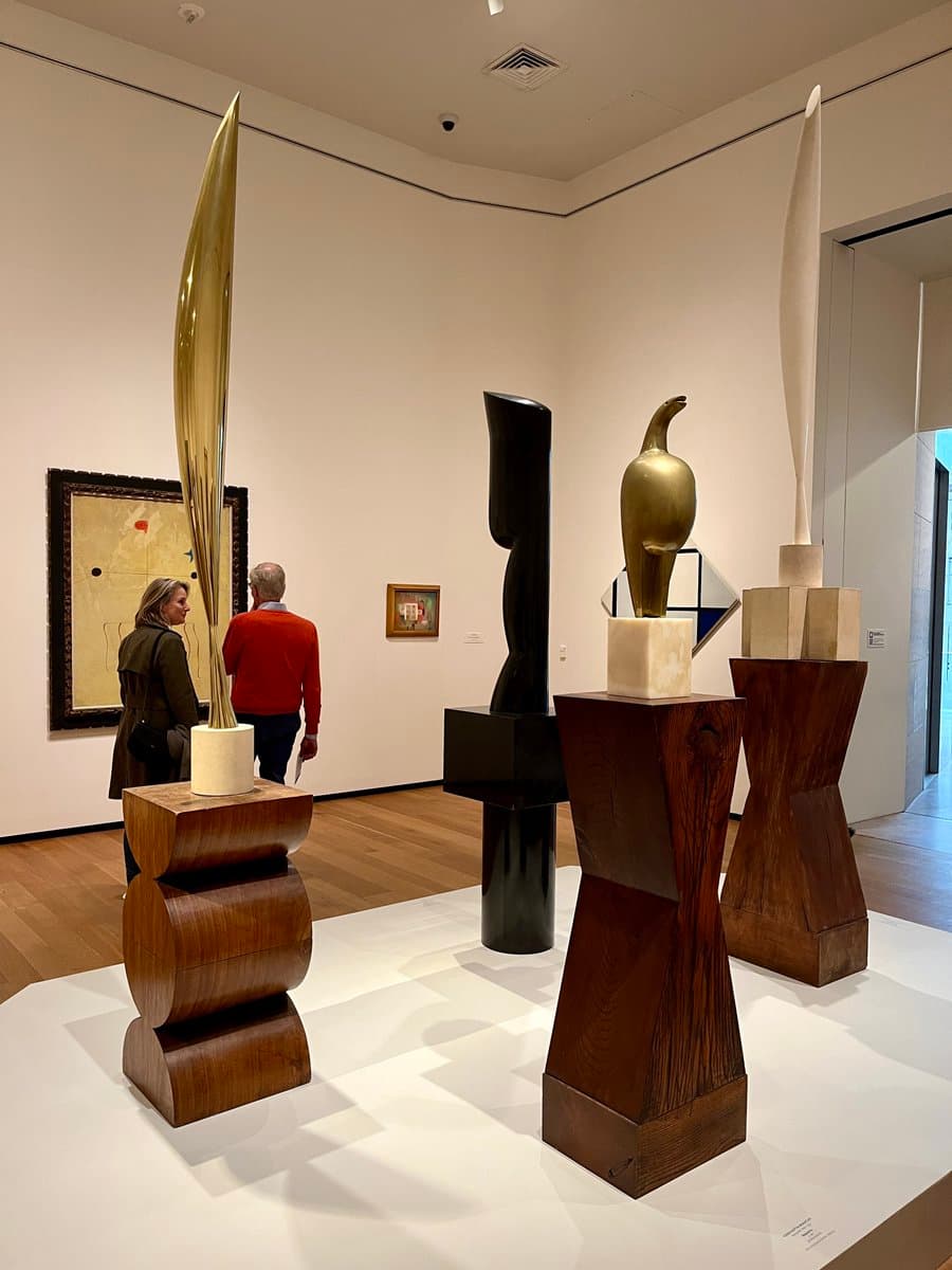 A photograph of a gallery exhibition featuring four tall, abstract sculptures on wooden pedestals, with two visitors viewing a framed painting on the wall in the background.
