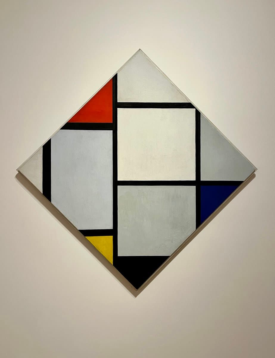 A photograph of a square canvas painting mounted on a wall and rotated 45 degrees to form a diamond shape. The artwork features a geometric, abstract composition in the style of Piet Mondrian, consisting of a grid of black lines dividing the canvas into blocks of white, light gray, red, yellow, and blue.