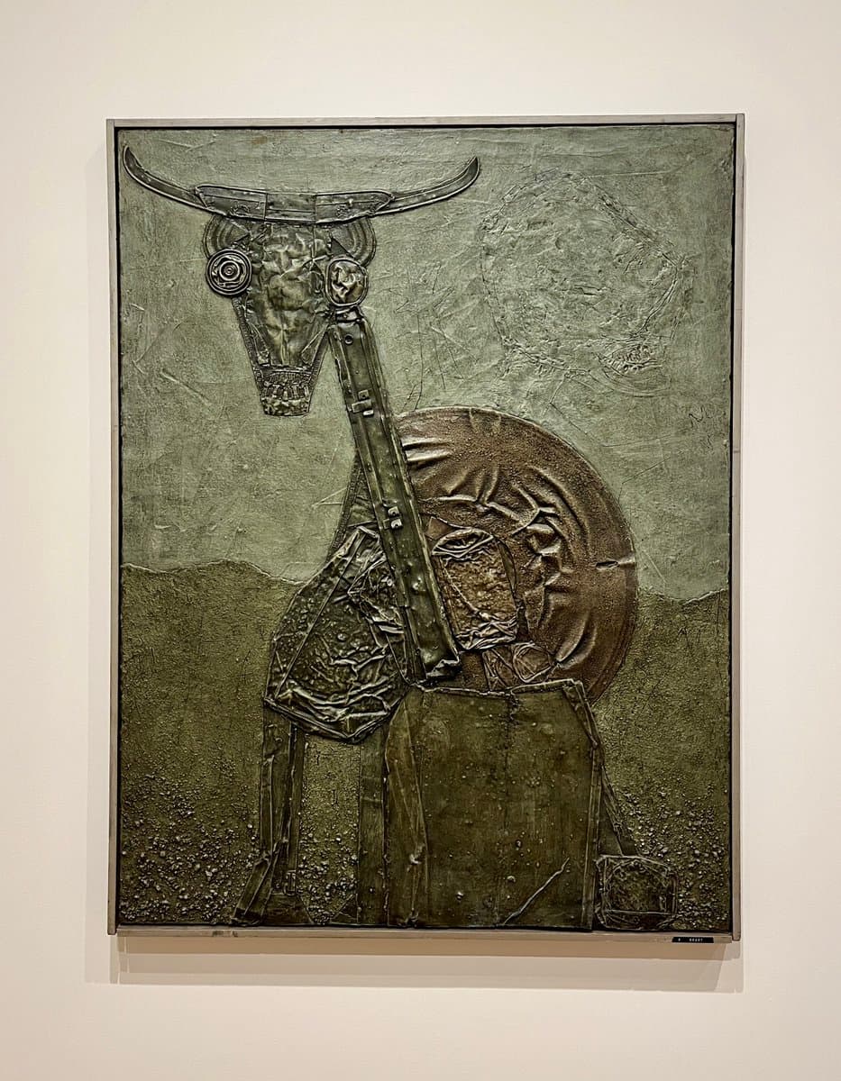 A photograph of a framed, textured metal relief artwork hanging on a white gallery wall. The piece depicts a stylized figure with a halo and a long, mechanical-looking neck extending up to a bull's skull with horns.