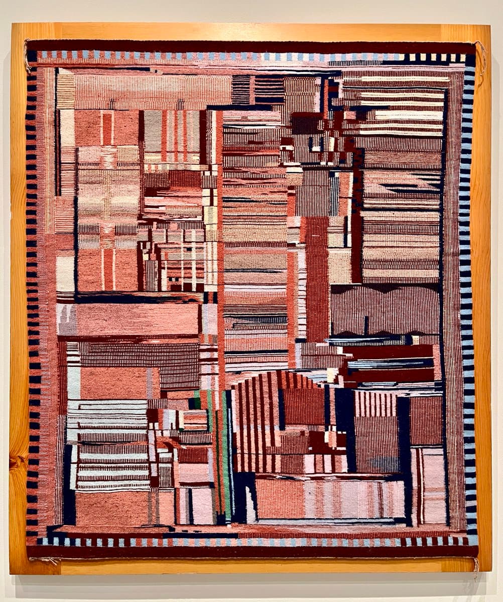 A framed textile artwork featuring an abstract, geometric pattern in shades of red, brown, and white, mounted on a wooden board.