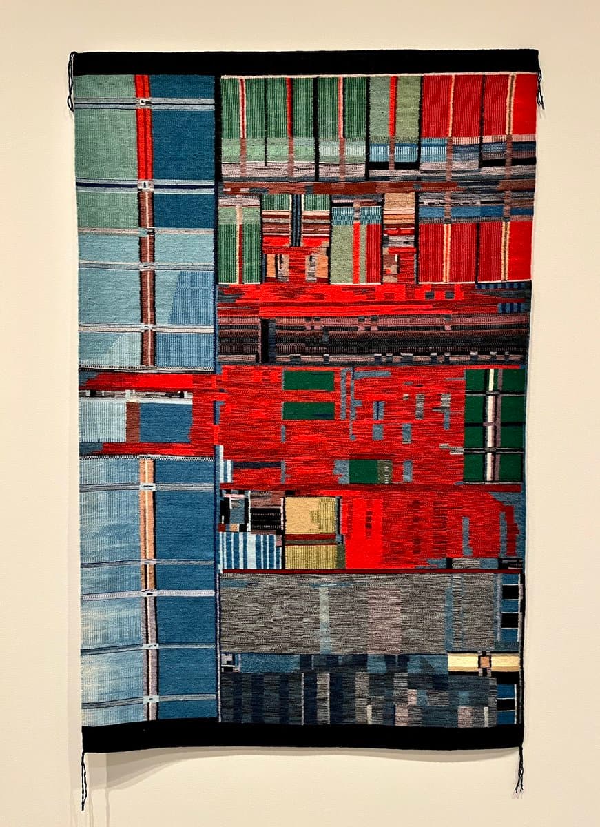 A vertical abstract woven textile hanging on a white wall, featuring a geometric patchwork of blue, red, green, and grey blocks.