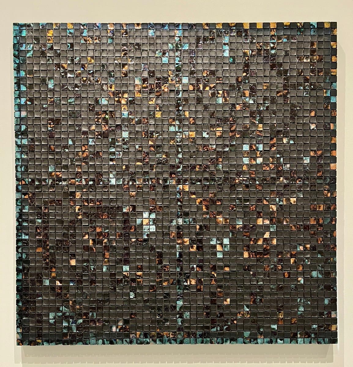 A square mosaic artwork composed of small, dark, iridescent tiles arranged in a grid pattern. The tiles reflect light in shades of blue, green, and gold, creating a shimmering, textured surface against a plain background.