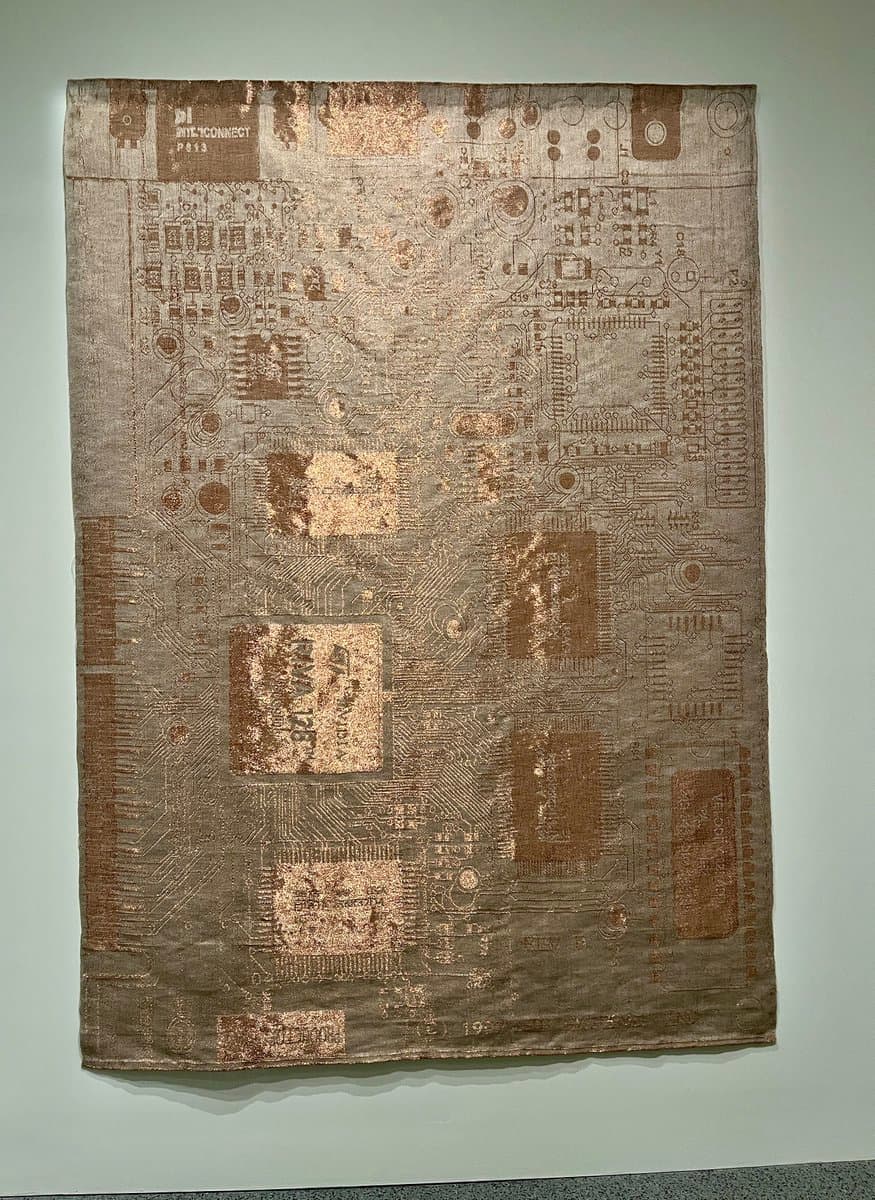 A photograph of a large, rectangular textile artwork hanging on a gallery wall, featuring a woven design that mimics the intricate circuitry of a computer motherboard. The piece is rendered in metallic bronze and silver threads, with visible text labels such as 'INTEL CONNECT' and 'RAM 128' integrated into the pattern.