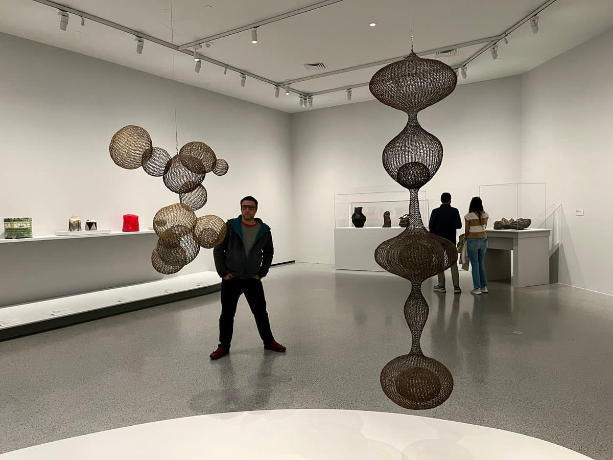 A wide shot of a museum gallery featuring large, suspended woven sculptures and visitors viewing the exhibits.