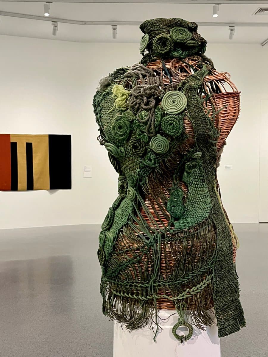 A photograph of a textile art installation in a gallery, featuring a wicker mannequin torso covered in intricate green woven and knotted fibers, including spiral and rosette patterns. In the background, a geometric textile piece with red, gold, and black vertical stripes hangs on the white wall.