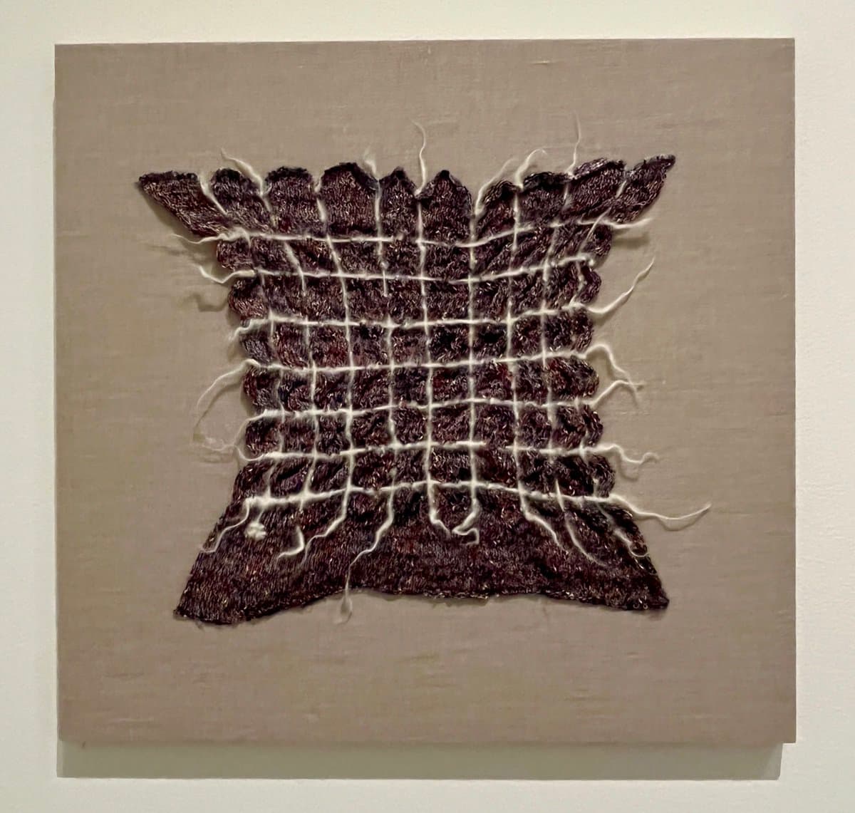 A photograph of a textile art piece featuring a dark, knitted or woven grid structure with loose white threads, mounted on a neutral beige background. The piece is displayed on a wall, likely in a gallery setting.