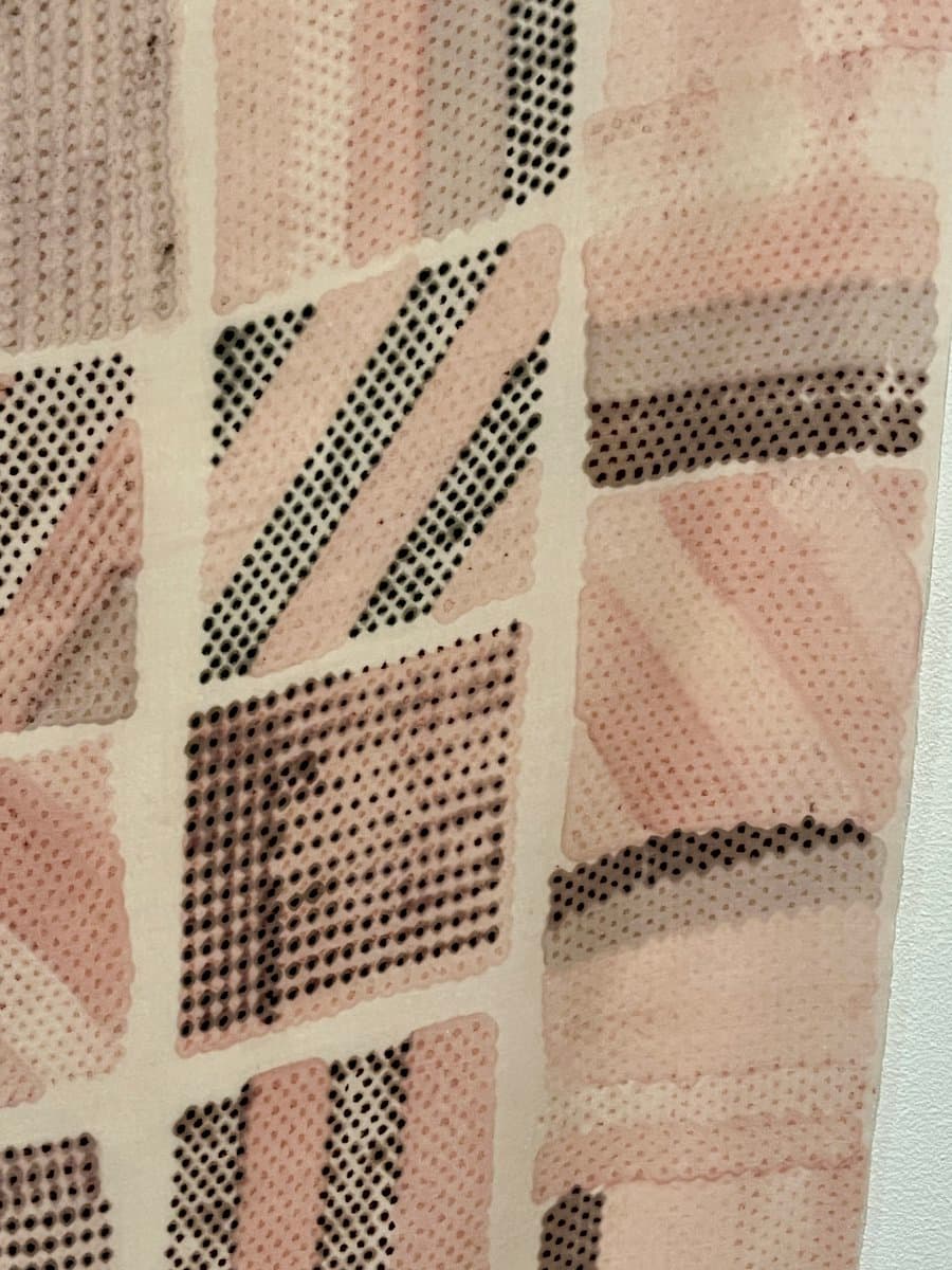 A close-up view of a textile artwork featuring a grid of rectangular panels in shades of pink, beige, and black. Each panel displays a distinct pattern of dots or stitches, creating a textured, abstract composition.
