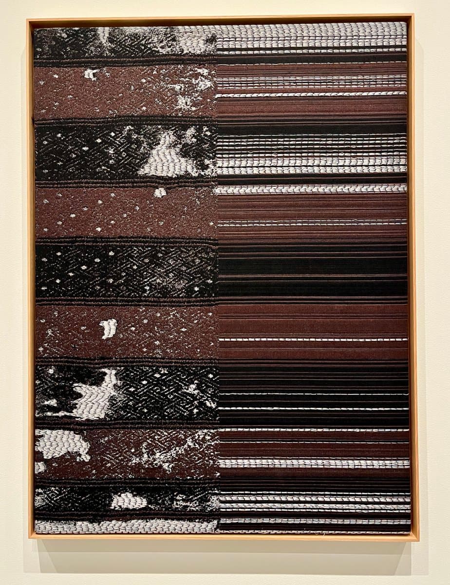 A framed textile artwork hanging on a white wall, featuring a grid of horizontal strips in black, white, and brown with varied woven textures and patterns. The piece is divided into two main sections: a left side with more abstract, mottled patterns and a right side with more uniform, striped lines.