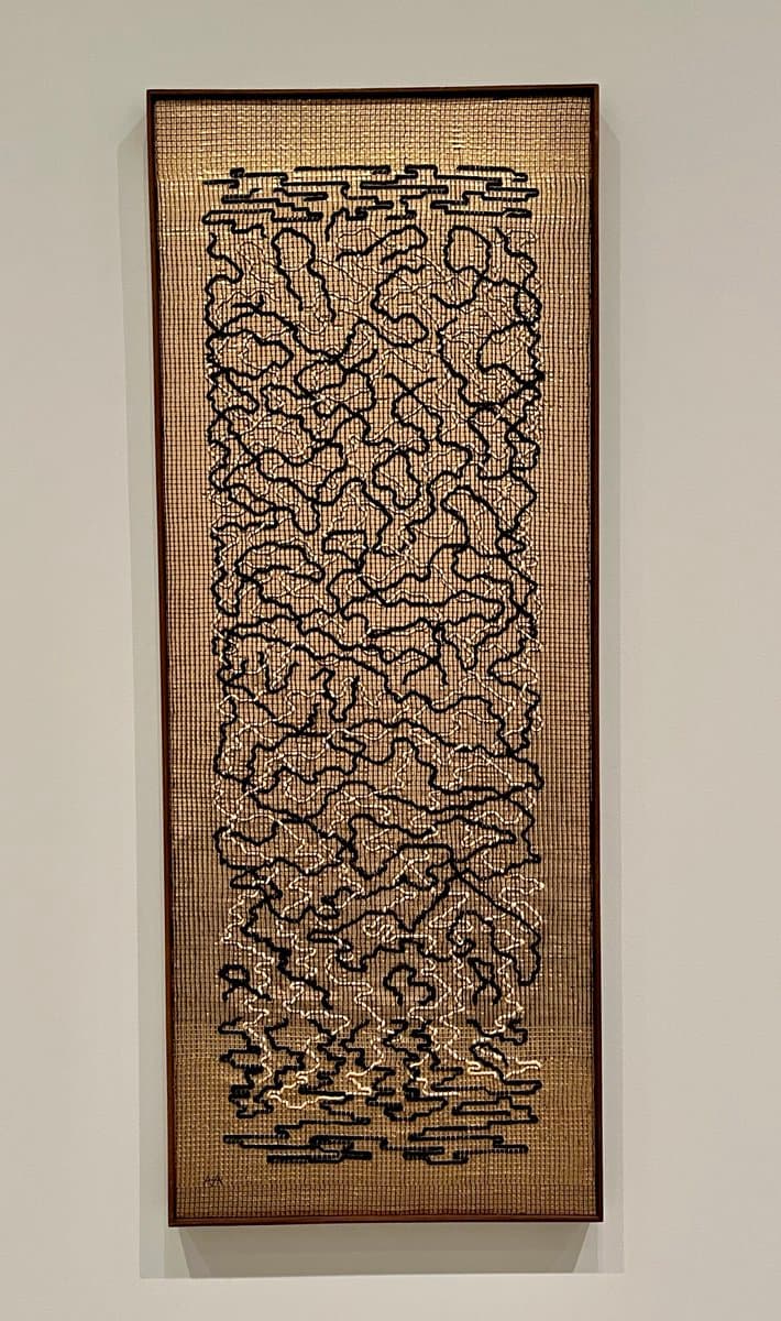 A tall, rectangular framed artwork hanging on a white wall, featuring a textured, woven background with an intricate black line drawing of abstract, cloud-like or organic forms. The piece is enclosed in a dark wooden frame and appears to be a textile or mixed-media art installation.