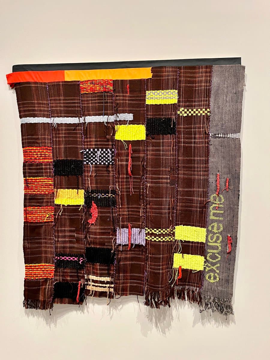 A photograph of a large, abstract textile artwork hanging on a white wall. The piece is composed of vertical strips of brown plaid fabric interspersed with patches of bright yellow, orange, black, and white woven material, featuring frayed edges and the word 'excuse me' embroidered vertically on the right side.