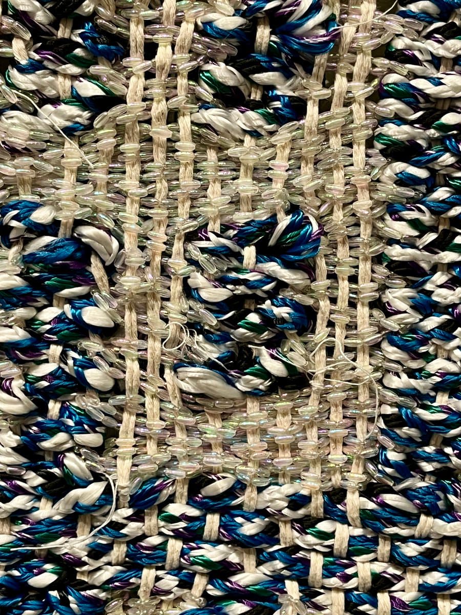 A close-up view of a woven textile featuring a complex pattern of white, blue, green, and purple threads interlaced with vertical strands of clear, iridescent beads. The fabric has a textured, handcrafted appearance with visible loose threads and a dense, intricate weave.