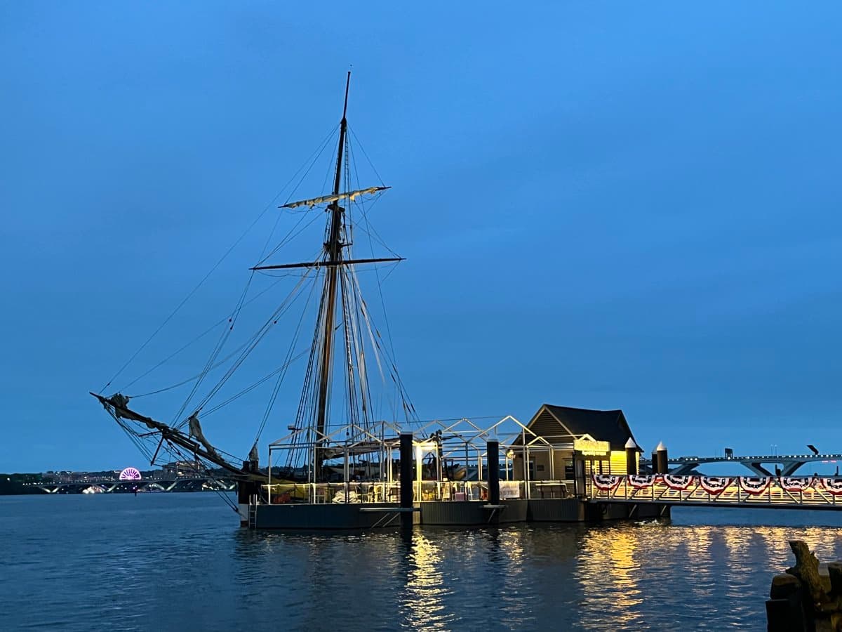 A tall-masted sailing ship is docked at a lit pier on a calm body of water during twilight, with a bridge and city lights visible in the background. The scene is illuminated by warm artificial lights from the dock and a nearby building, reflecting on the water's surface.