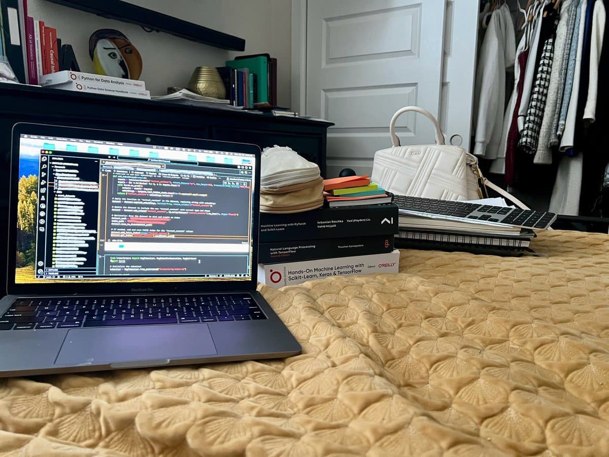 A laptop displaying a code editor sits on a bed next to a stack of machine learning textbooks, a white handbag, and a keyboard, with a closet visible in the background. The scene depicts a casual workspace set up on a bed.