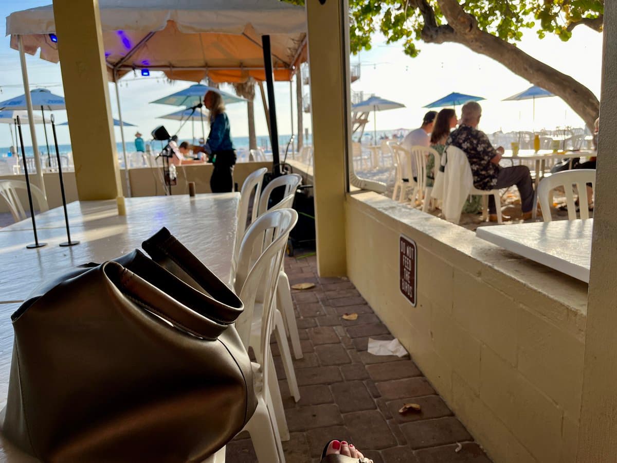 A first-person view from a beachside restaurant patio, showing a brown leather handbag on a table in the foreground and a view of the ocean and other patrons through the open-air structure. A small sign on the wall reads "DO NOT FEED THE HIPPOS." The scene includes white plastic chairs, a brick floor, and a person's foot with red nail polish visible at the bottom.