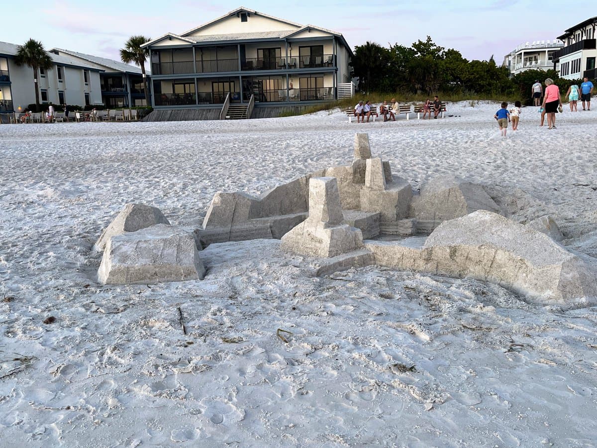 A wide shot of a sandy beach featuring a large, intricately carved sand sculpture in the foreground, with beachgoers and multi-story coastal buildings in the background.