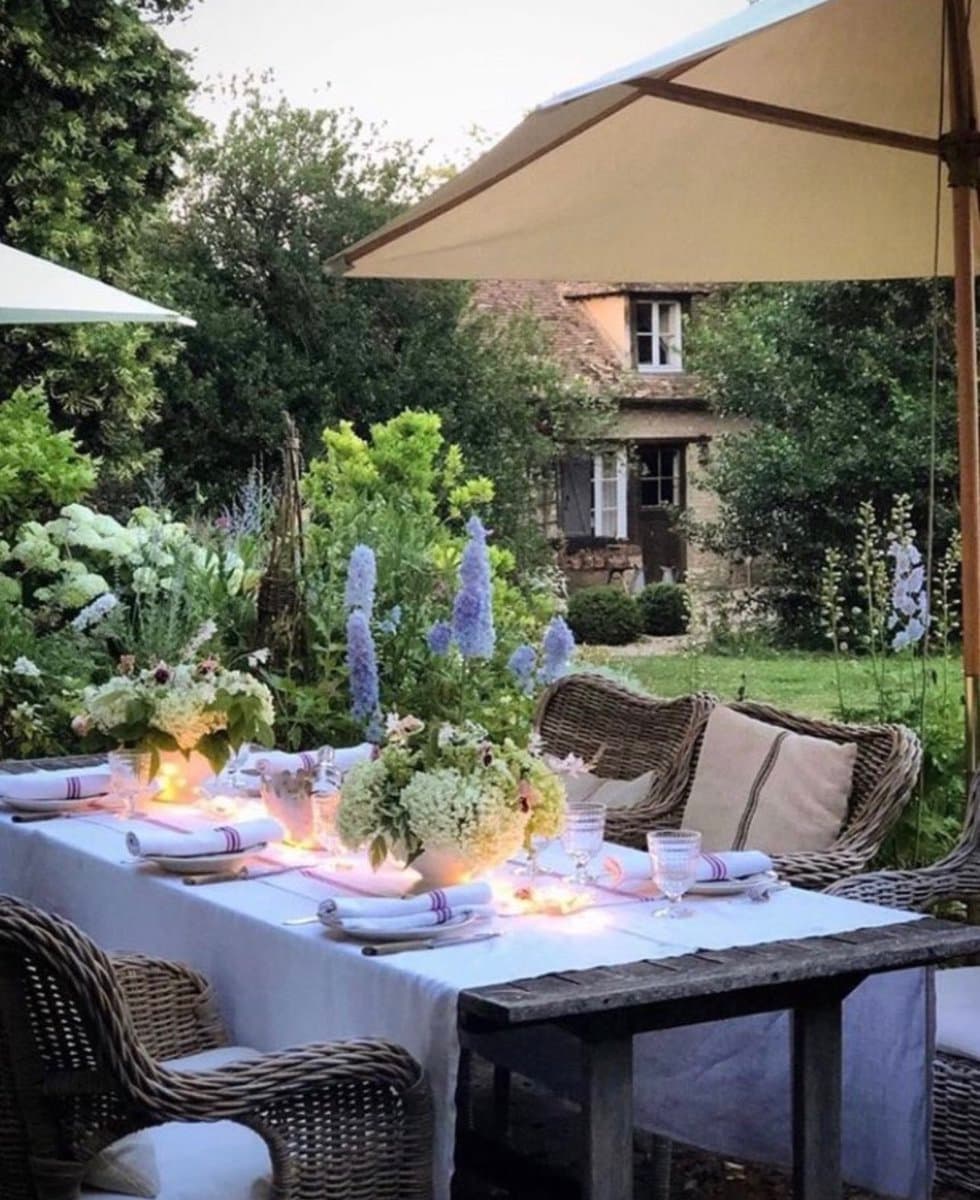 A photograph of an outdoor dining table set for a meal in a lush garden, featuring white linens, floral centerpieces, and candles under a large umbrella. In the background, a stone house is visible among the greenery.