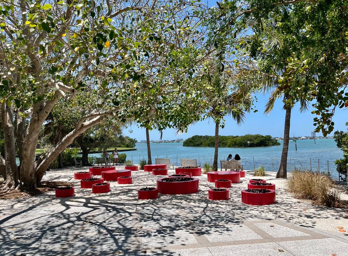 A sunny outdoor scene at a waterfront park, featuring a paved area with numerous red circular planters and benches under the shade of large trees. In the background, a body of water with a small island and distant buildings is visible.