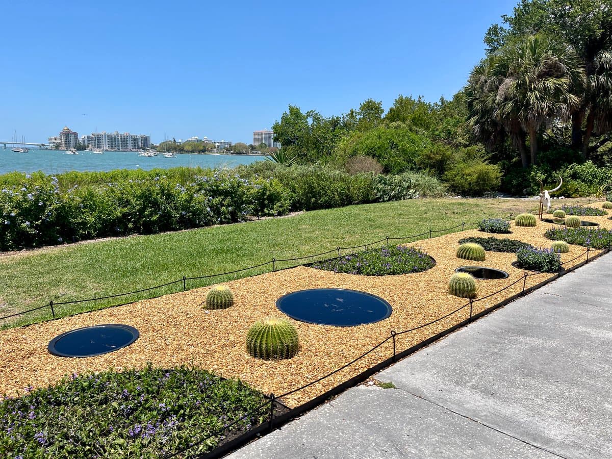 A sunny outdoor view of a landscaped garden featuring a gravel bed with barrel cacti and circular black mirrors, bordered by a low fence and a concrete walkway. In the background, there is a grassy area, lush greenery, and a body of water with boats and a city skyline visible in the distance.
