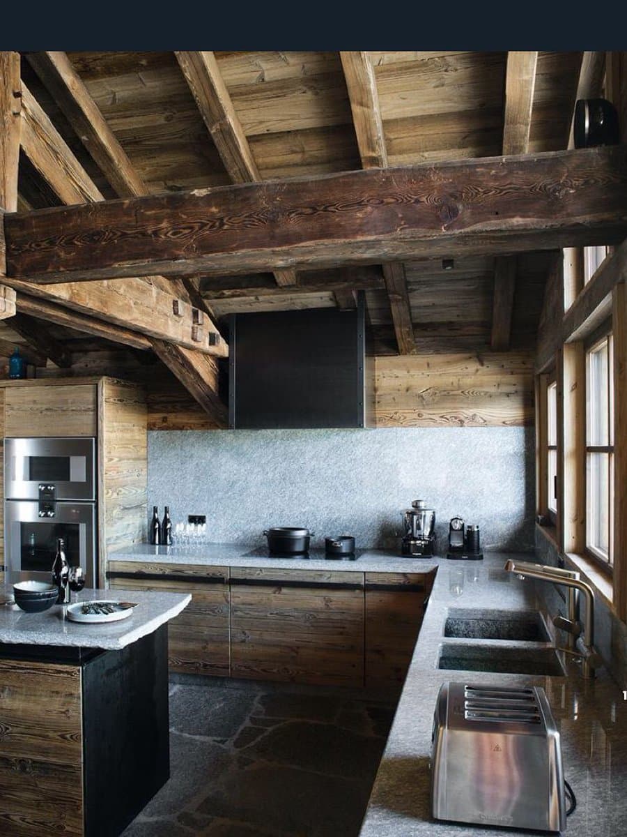 A rustic kitchen interior featuring exposed wooden ceiling beams, wood-paneled walls, and stone countertops. The space includes a large island, built-in stainless steel appliances, and a farmhouse-style sink.