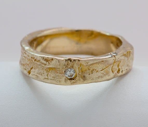 A close-up photograph of a textured, hammered gold ring featuring a small, round diamond set into its front face. The ring is displayed against a plain white background.
