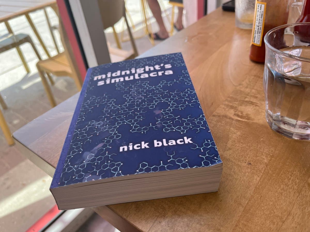A paperback book titled 'midnight's simulacra' by 'nick black' rests on a wooden table next to a glass of water. The book has a dark blue cover featuring a white molecular structure pattern.