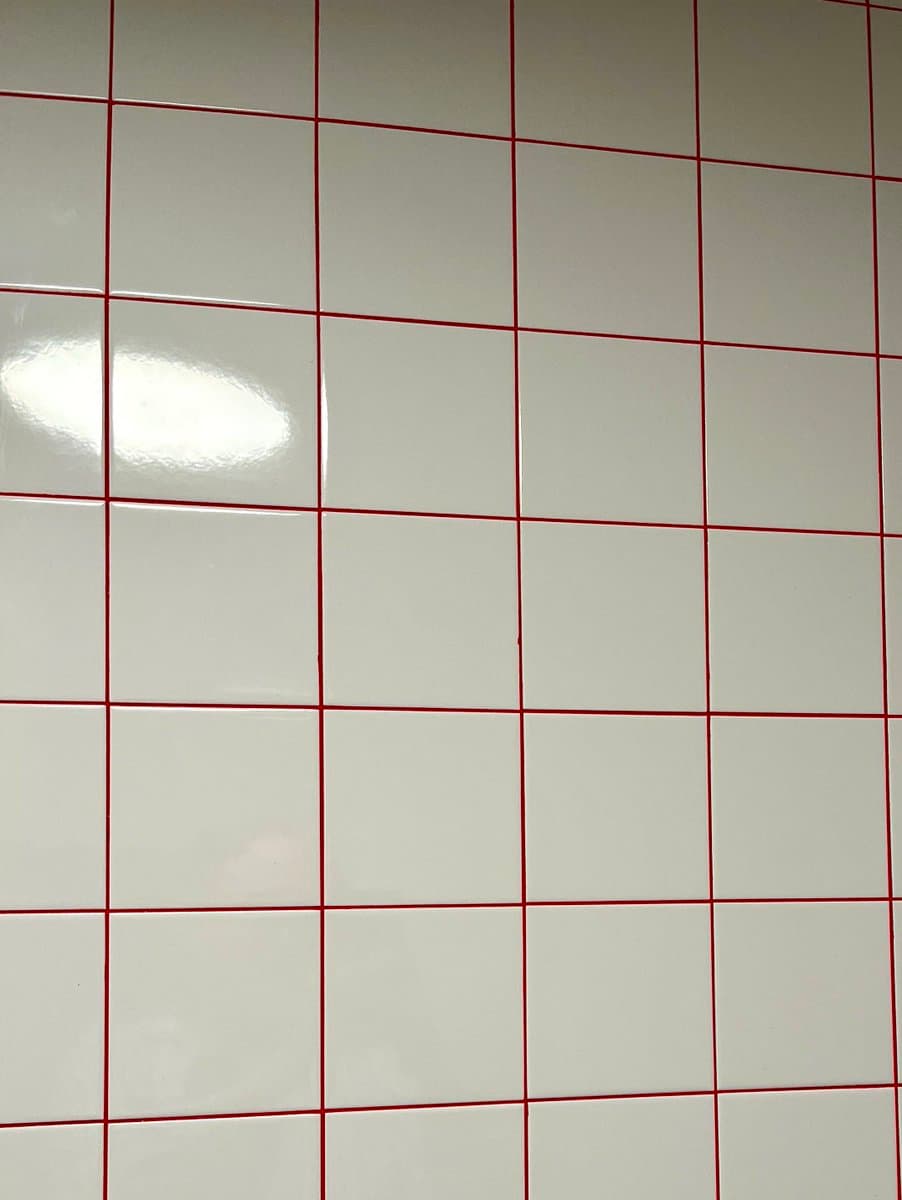A close-up view of a wall covered in glossy white square tiles separated by thin, bright red grout lines. A bright light reflection is visible on the tiles in the upper left area.