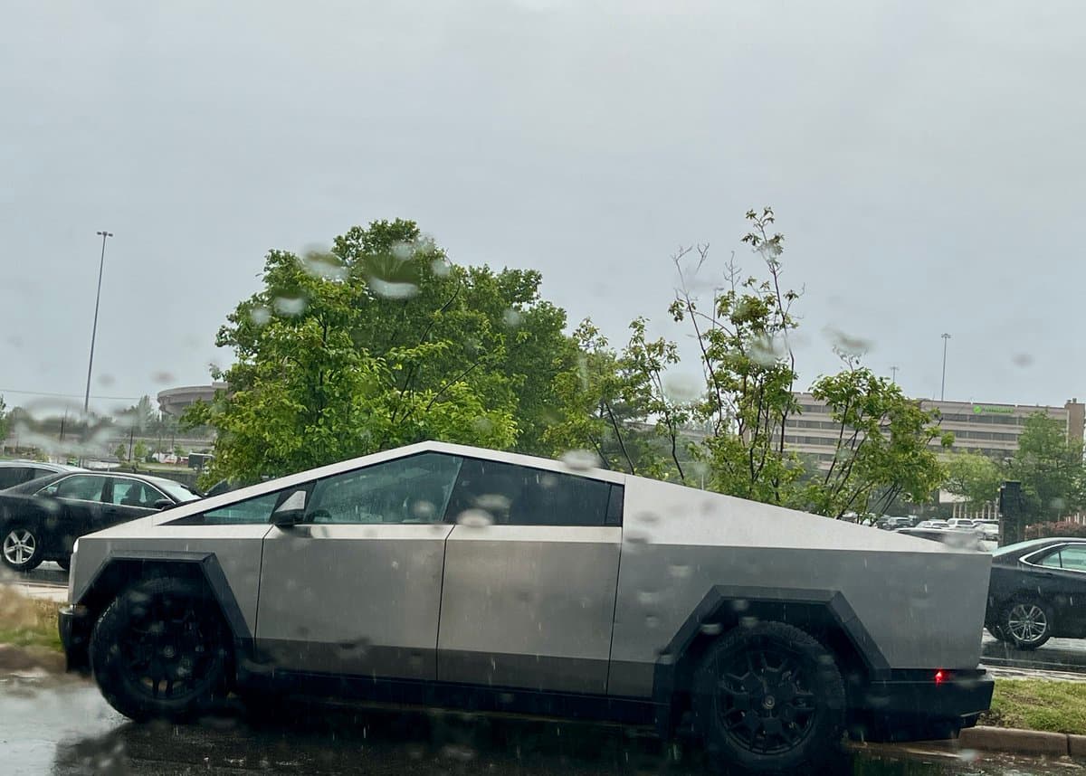 A photograph of a silver Tesla Cybertruck parked in a wet parking lot on a rainy day, viewed through a window with water droplets on the glass.