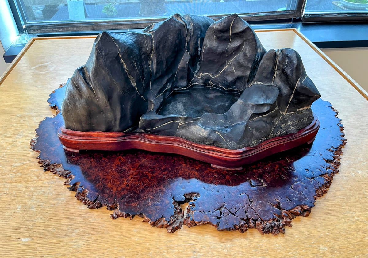 A large, dark, sculpted stone with a mountain-like shape sits on a polished wooden base, which rests on a decorative, irregularly shaped wooden slab on a table. The scene is set indoors near a window, with the stone sculpture being the central focus.