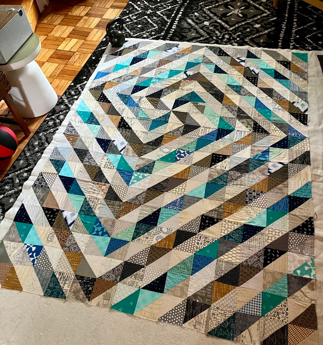 A top-down view of a large, in-progress patchwork quilt featuring a geometric pattern of triangles in shades of blue, teal, brown, and white, laid out on a patterned rug.