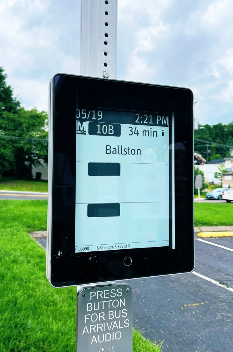 A close-up photograph of an e-ink bus status sign mounted on a metal pole at a bus stop, displaying the date, time, and next bus arrival information. Below the screen is a small metal plaque with instructions for an audio feature.
