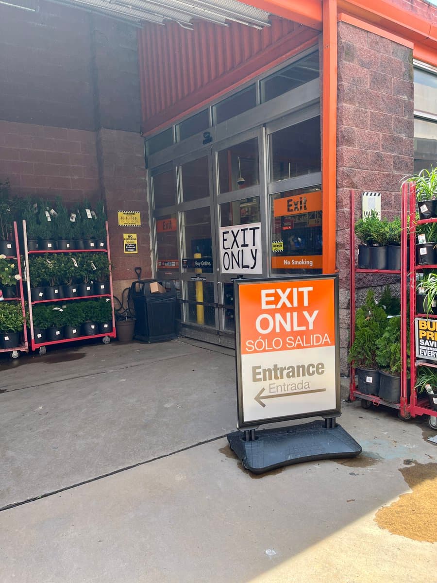 A storefront entrance with multiple signs indicating it is an exit-only area, including a large orange and white A-frame sign on the ground and smaller signs on the glass doors. Potted plants are displayed on red racks on either side of the entrance.