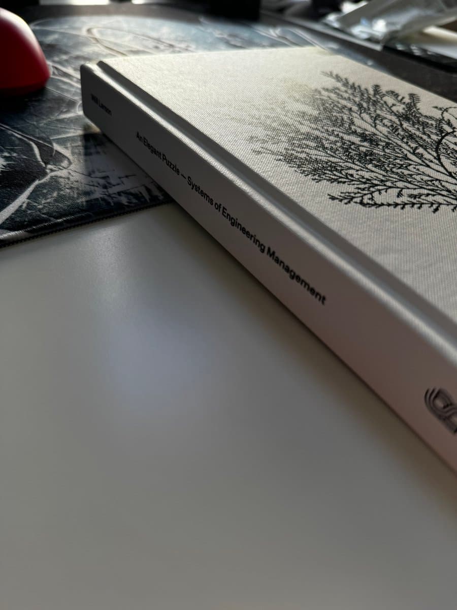 A close-up photograph of a hardcover book resting on a desk, showing its spine and part of its cover. The book has a white spine with black text and a textured, off-white cover featuring a black botanical illustration.