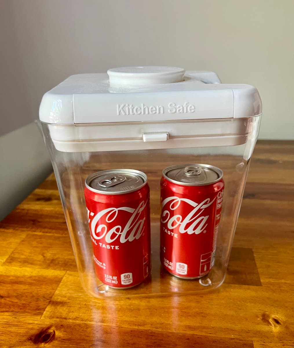 A clear plastic container labeled "Kitchen Safe" sits on a wooden table, holding two red cans of Coca-Cola inside. The container has a white lid and is positioned against a plain wall.