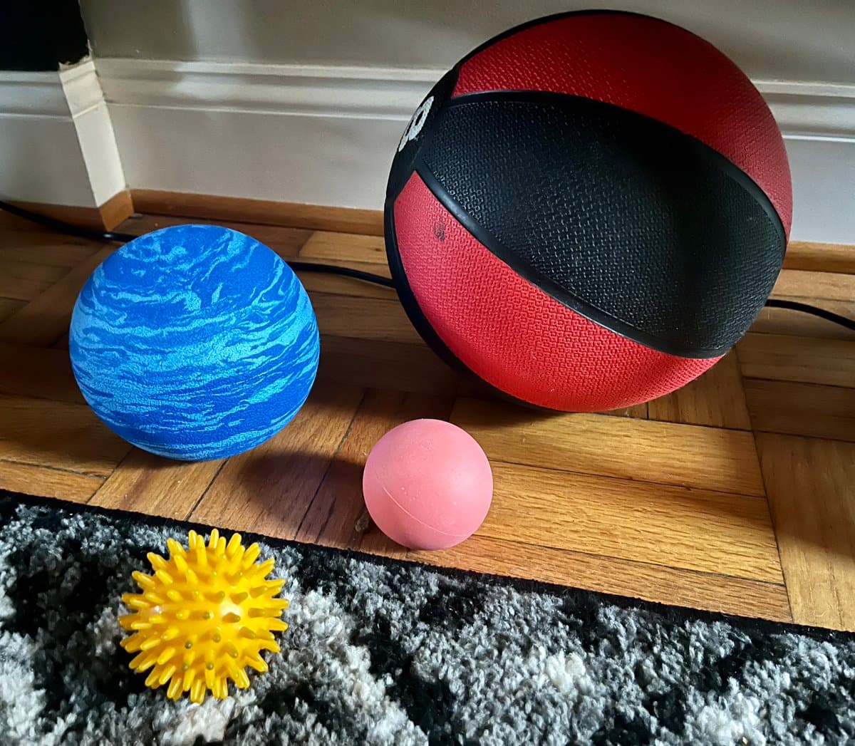 A collection of four balls of varying sizes and textures arranged on a wooden floor and a black-and-white rug. The group includes a large red and black basketball, a medium-sized blue ball with a marbled pattern, a small pink ball, and a small yellow spiky ball.