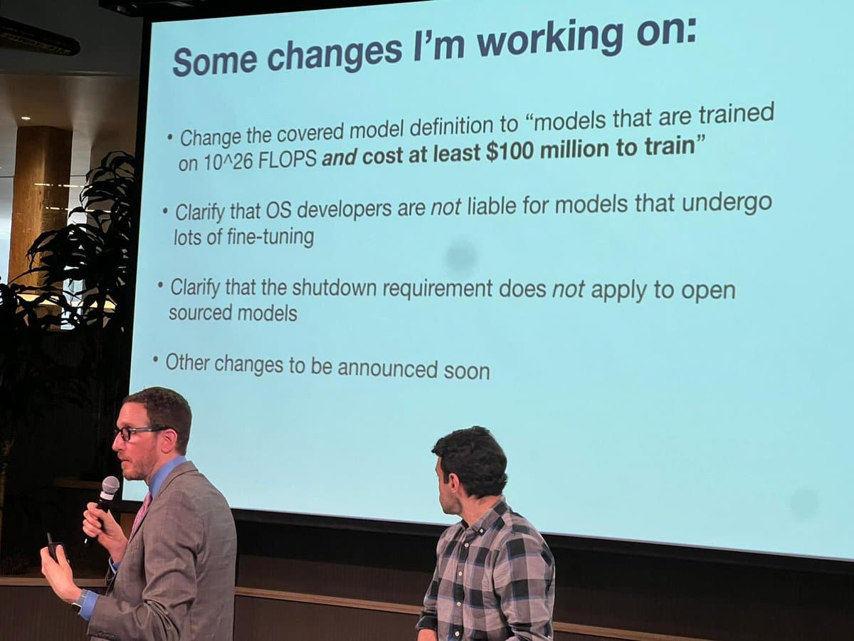 A photograph of a presentation at a conference, showing two men on stage in front of a large projection screen. The screen displays a slide titled "Some changes I'm working on:" with bullet points regarding model definitions and liability.