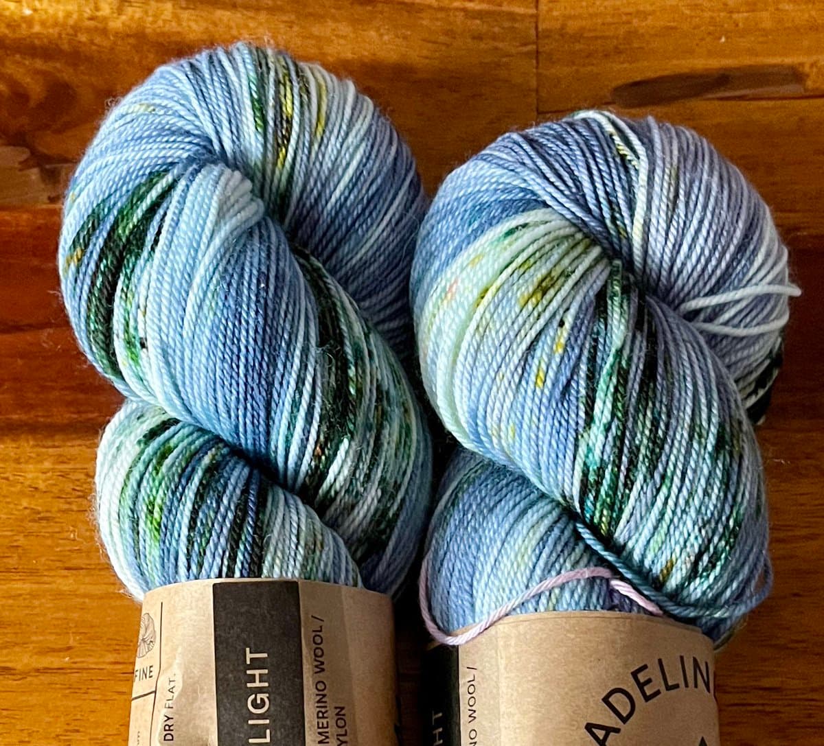 Two skeins of variegated blue, green, and white yarn resting on a wooden surface. The paper labels on the skeins indicate the brand is Madelinetosh and the weight is Light.