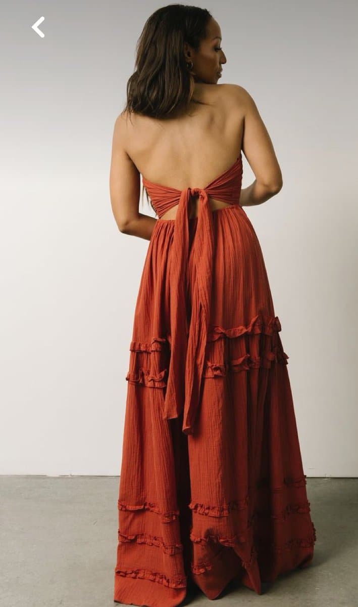 A woman is shown from behind, wearing a long, rust-colored sundress with a tie-back detail and ruffled tiers on the skirt. The dress features a crinkled texture and a low back, and she is standing against a plain white wall.