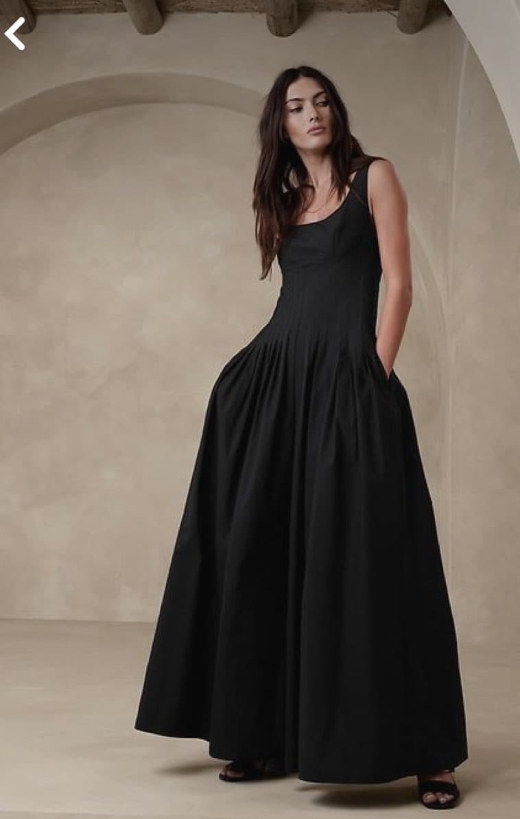 A woman with long dark hair stands in a minimalist, beige-toned room wearing a long, black sleeveless sundress with a pleated skirt. She is posing with one hand in her pocket, looking slightly away from the camera. The image appears to be a fashion product photo, likely from a Pinterest search result for 'sundress'.
