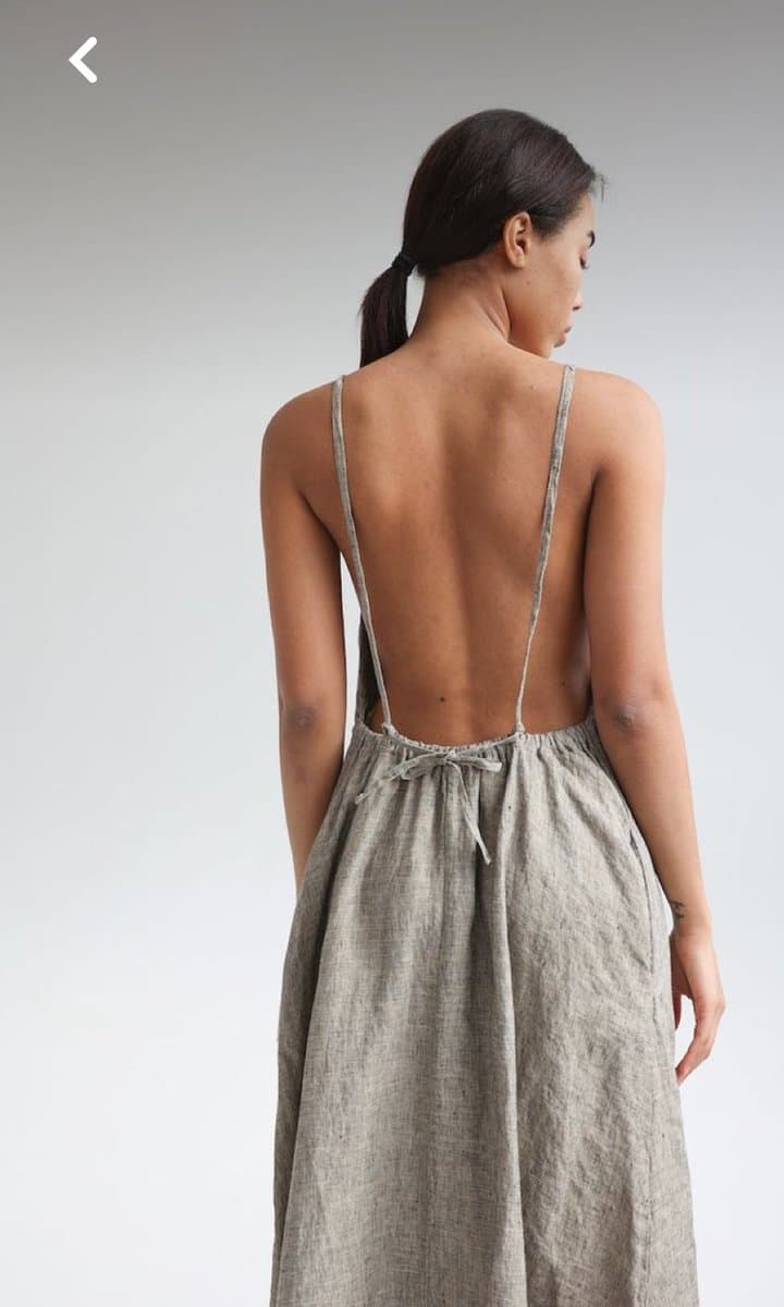 A woman with dark hair in a ponytail is shown from behind, wearing a light gray, open-back dress with thin straps and a tie at the waist. The image has a plain white background and a small white arrow icon in the upper left corner, suggesting it is a screenshot from a mobile app or website.