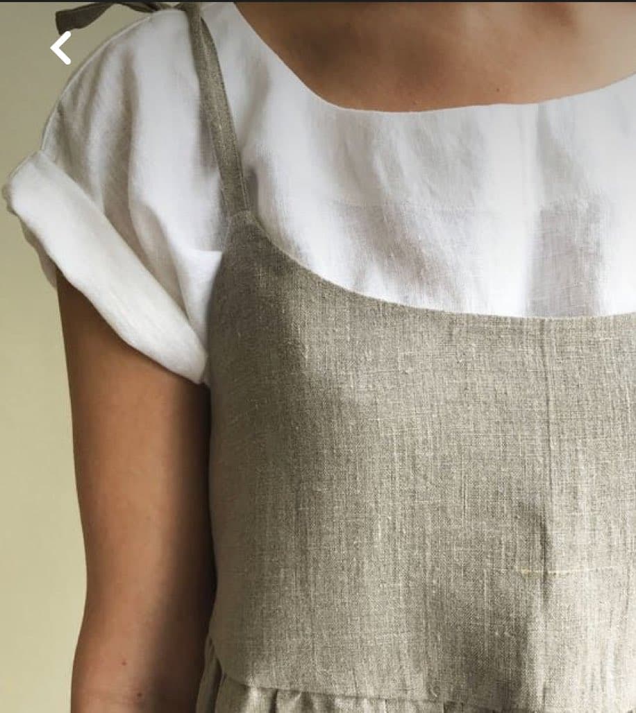 A close-up photograph of a person's torso wearing a white short-sleeved shirt underneath a textured, light brown linen apron or pinafore dress. The image focuses on the fabric textures and the garment's straps.
