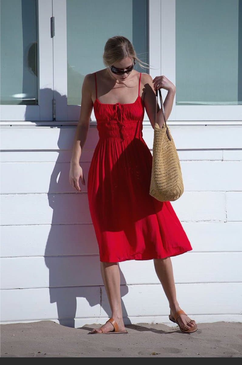 A woman wearing a red midi dress, sunglasses, and sandals stands against a white wall while holding a woven bag.