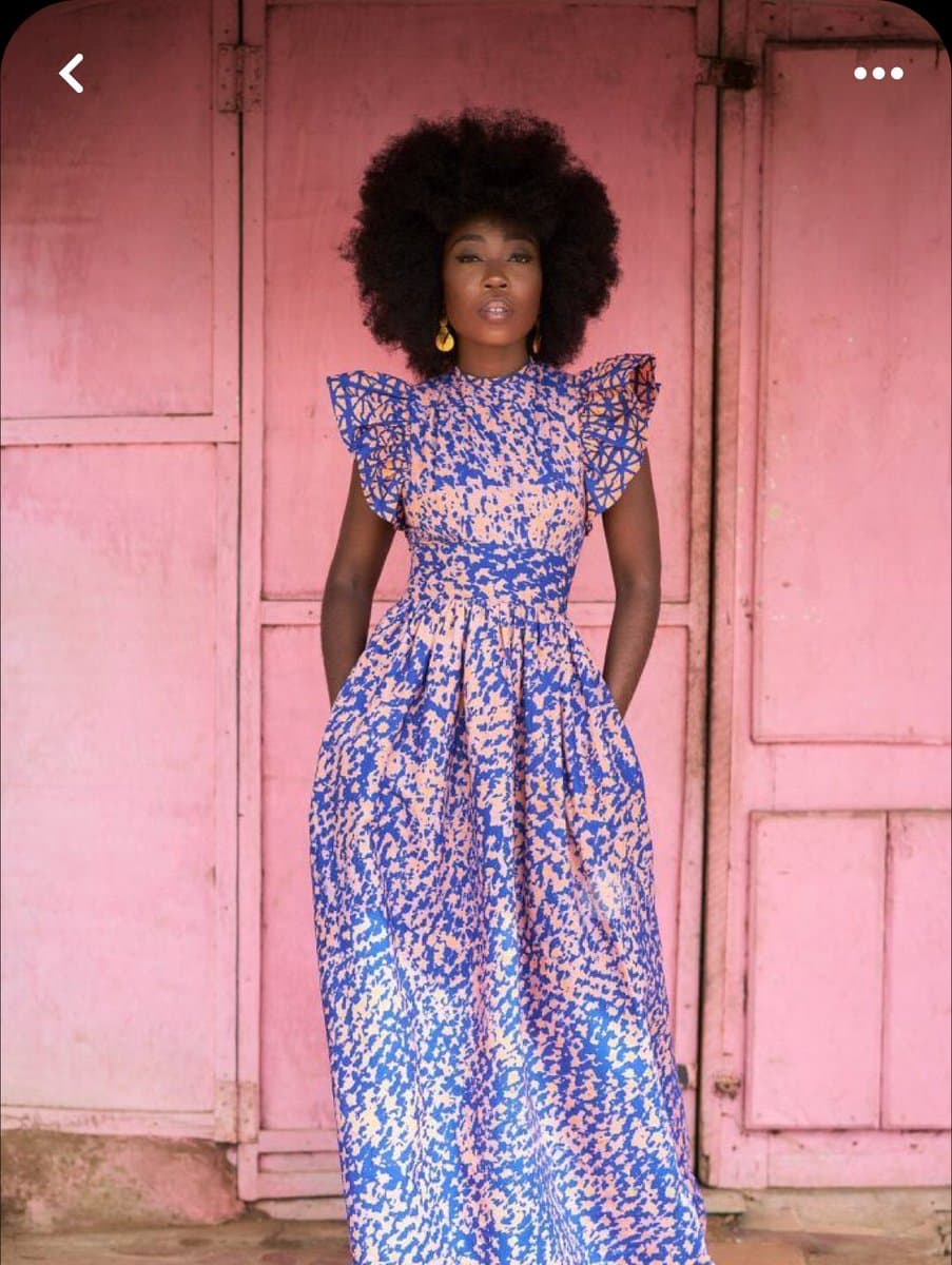 A woman with a large afro hairstyle stands in front of a pink wooden door, wearing a long, patterned blue and pink dress with ruffled sleeves. She is posing for a portrait with her hands in her pockets.