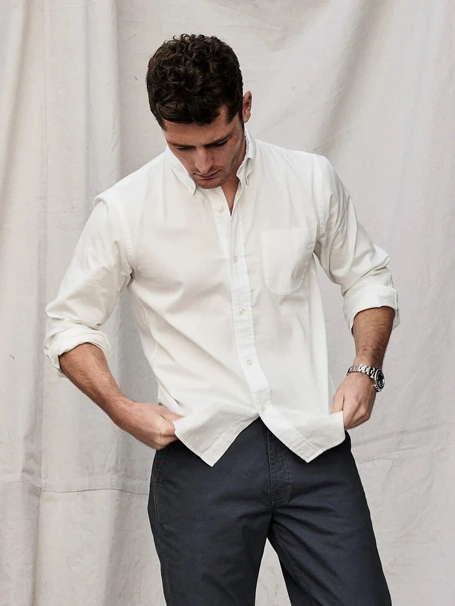 A man with short dark hair stands in front of a white backdrop, wearing a white button-down shirt with the sleeves rolled up to his elbows and dark trousers. He is looking down while pulling the bottom hem of his shirt with both hands, and he wears a silver wristwatch on his left wrist.