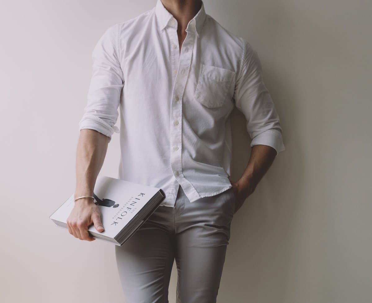 A person wearing a white button-down shirt with rolled-up sleeves and light gray pants, holding a book titled 'KINFOLK' against a plain wall.