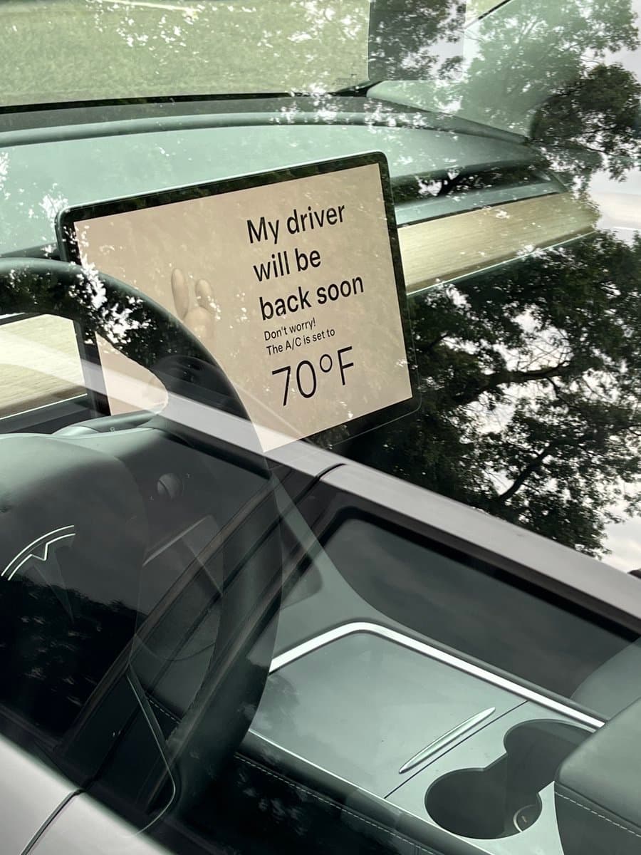 A photograph taken through the windshield of a Tesla, showing the central dashboard screen displaying a message to a pet left in the car. The screen reads, "My driver will be back soon. Don't worry! The A/C is set to 70°F." The car's interior, including the steering wheel and center console, is visible, with reflections of trees on the glass.